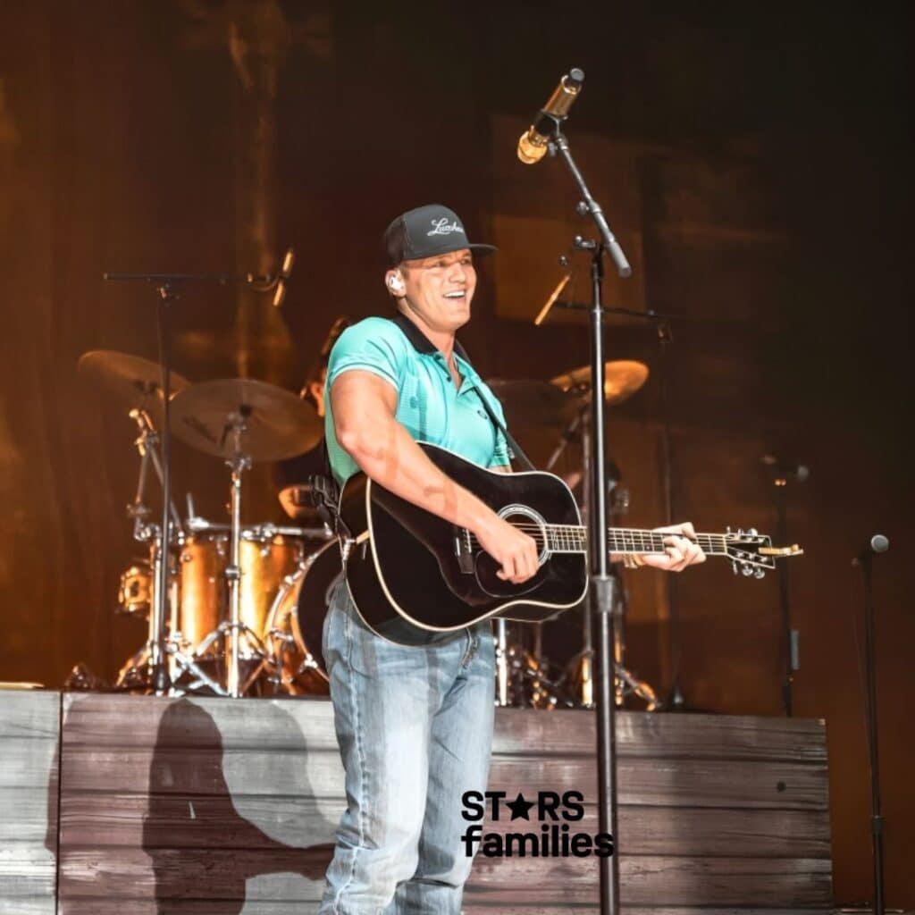 Parker McCollum, wearing a light blue shirt, jeans, and a black cap, stands on stage holding an acoustic guitar. He is in front of a microphone. Behind him, there is a drum set and other musical equipment. The background is dark with warm stage lighting.