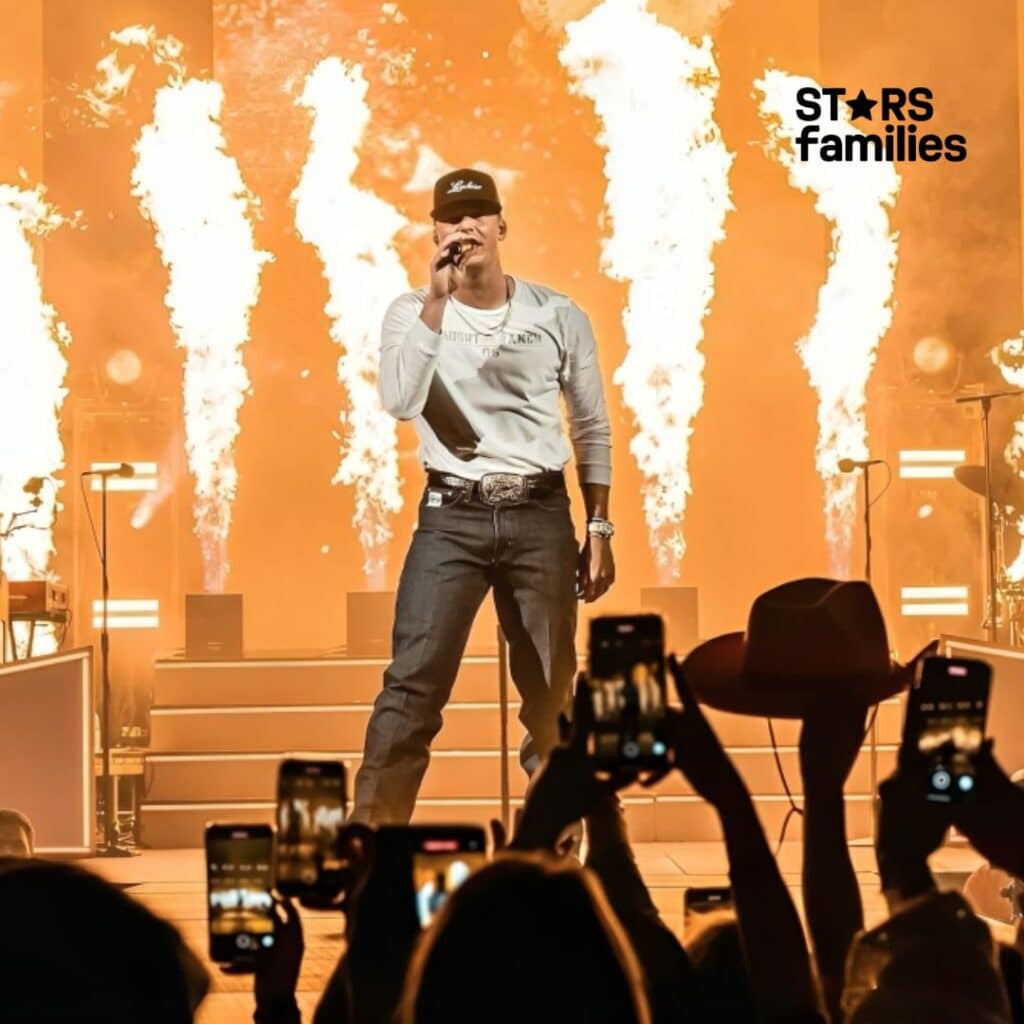 Parker McCollum performs on stage wearing a white long-sleeve shirt, dark jeans, a belt with a large buckle, and a black cap. The background features large flames and bright lights, creating a dramatic and energetic atmosphere. The audience in the foreground holds up smartphones, capturing the performance.