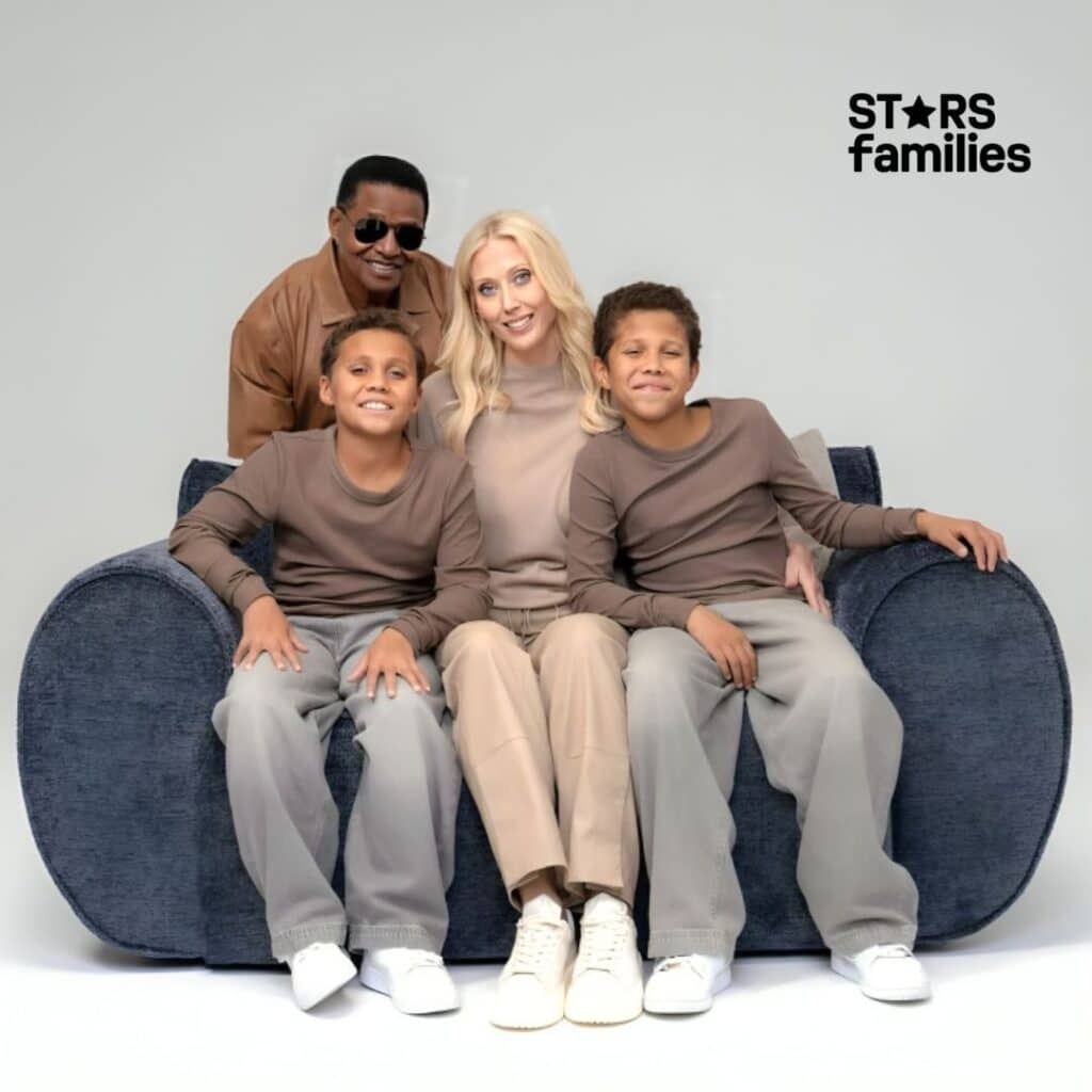 Jackie Jackson and his wife are sitting on a blue couch, with their twin sons seated on either side of them. Jackie, wearing neutral-toned clothing, sits on the left, while his wife, also dressed in neutral tones, stands behind the couch. The twin sons are wearing matching gray pants and brown long-sleeve shirts.