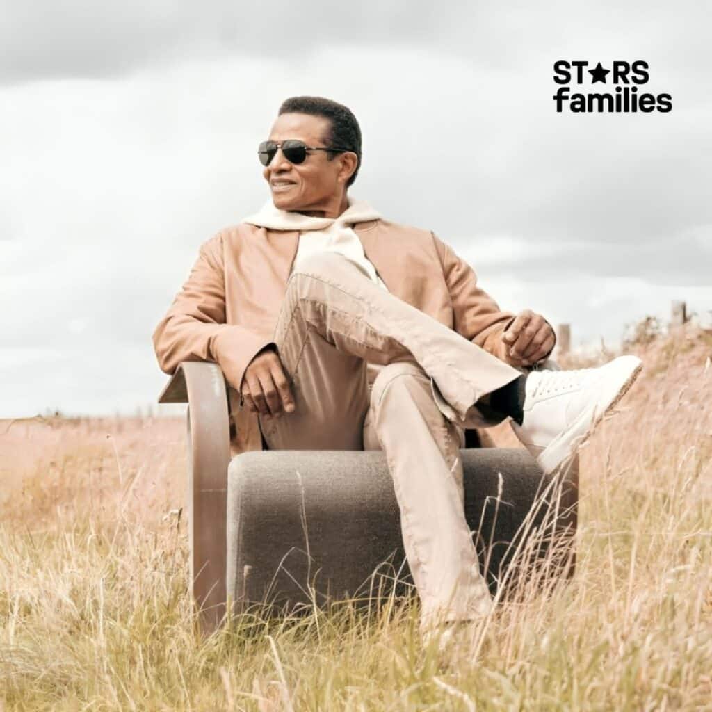 In a serene outdoor setting, Jackie Jackson, dressed in a light brown jacket, beige pants, and white sneakers, sits on a chair in a field of tall grass.