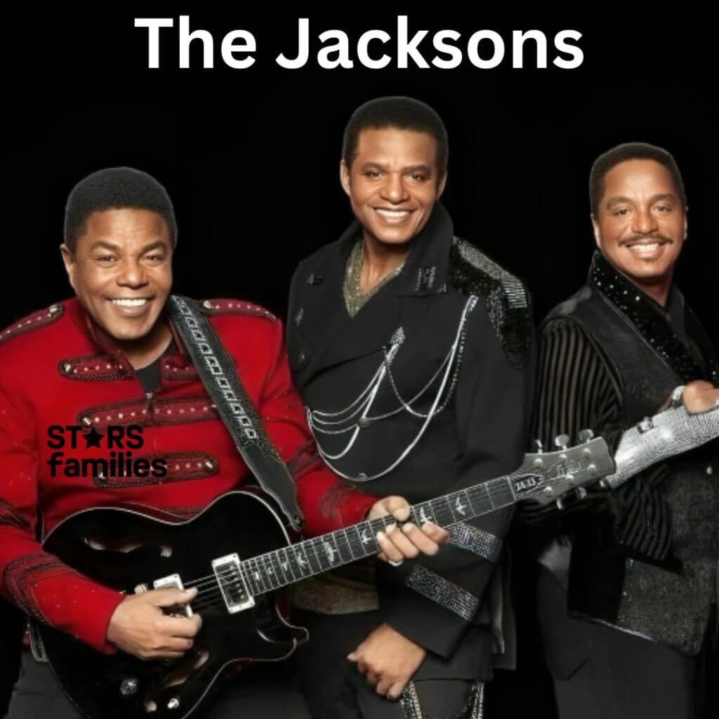 Three members of The Jacksons, dressed in elaborate, stylish outfits, pose together. The person on the left wears a red jacket with decorative elements and holds a black electric guitar.