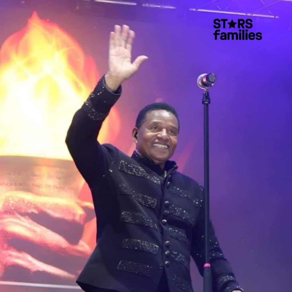 Jackie Jackson, wearing a black jacket with glittery embellishments, stands on stage with a microphone stand, raising one hand.