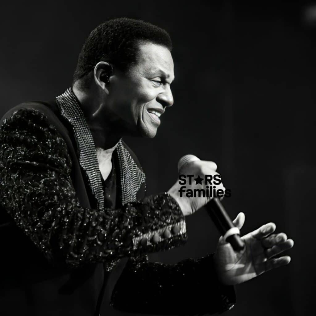 In a live performance setting, Jackie Jackson, dressed in a dark sequined jacket with a patterned collar, holds a microphone and sings on stage. The spotlight emphasizes his detailed jacket and the microphone, contrasting with the dark background.