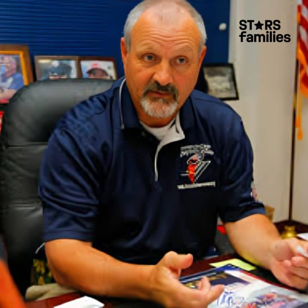 : Frank Siller, wearing a navy blue polo shirt with a logo on the left chest area, is seated at a desk. The desk has various items, including papers, a pen, and a framed photograph. The background features more framed photographs and an American flag.