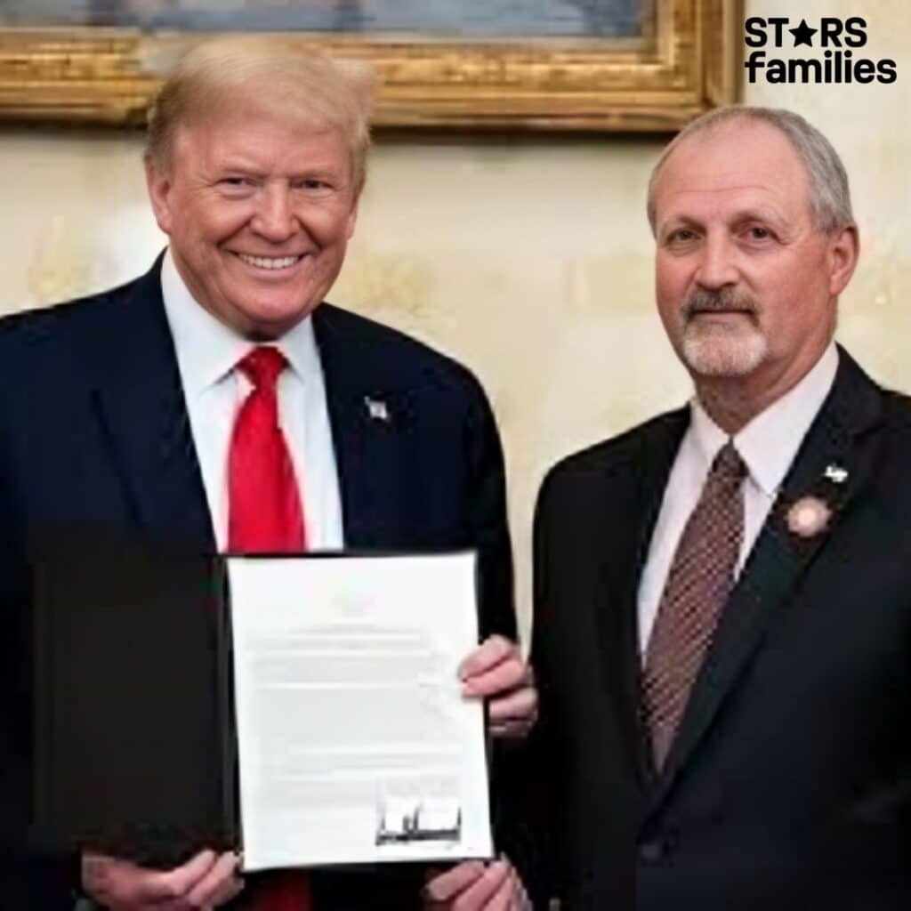In a formal setting, Frank Siller, dressed in a dark suit with a white shirt and a patterned tie, stands beside Donald Trump, who is wearing a dark suit with a white shirt and a red tie. Frank Siller holds a document in a black folder.