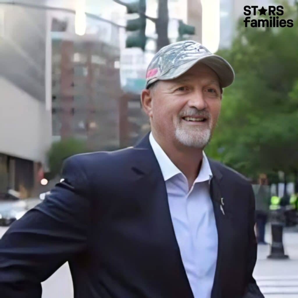 Frank Siller, wearing a dark suit jacket, a white dress shirt, and a camouflage baseball cap, stands in an urban setting with tall buildings, trees, and traffic lights in the background.