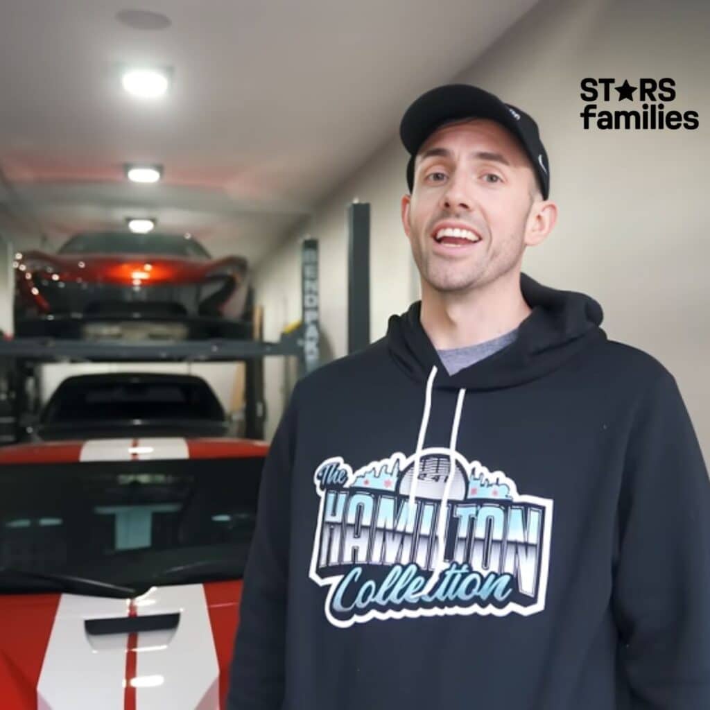 Steve Hamilton, wearing a black hoodie with the text "The Hamilton Collection" printed on it, stands in a garage. Behind him, there are two sports cars; one is red and positioned on a car lift, while the other is below it. The garage also has various automotive equipment and tools.