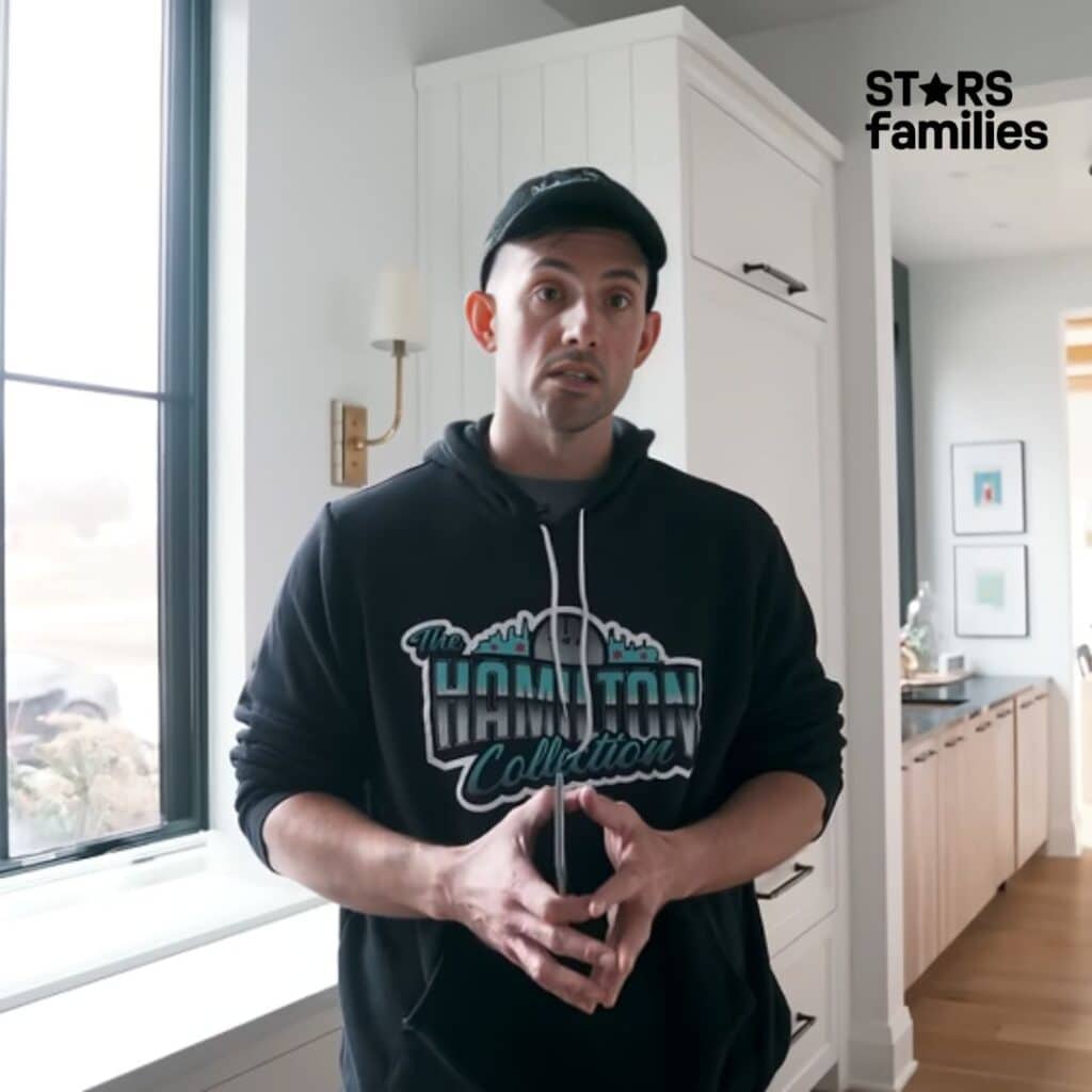 Steve Hamilton, wearing a black hoodie with the text "The Hamilton Collection" and a graphic of a city skyline, stands in a modern kitchen. He is also wearing a black cap. The background includes a large window, a wall-mounted light fixture, and a white cabinet.