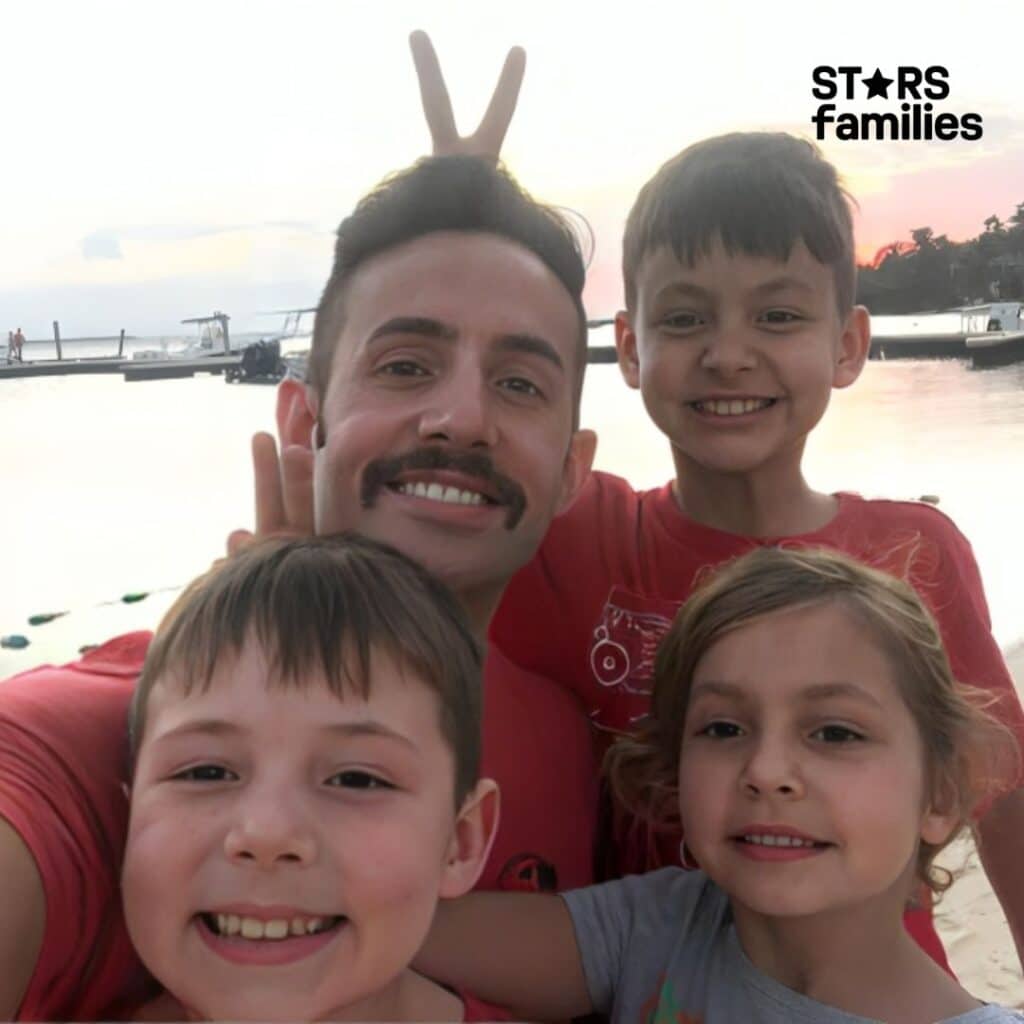 Steve Hamilton, wearing a blue plaid shirt and jeans, stands on a beach at sunset with his children. One of the children playfully makes a "bunny ears" gesture behind Steve's head. The background includes a calm body of water with boats docked at a pier and a sandy shore.