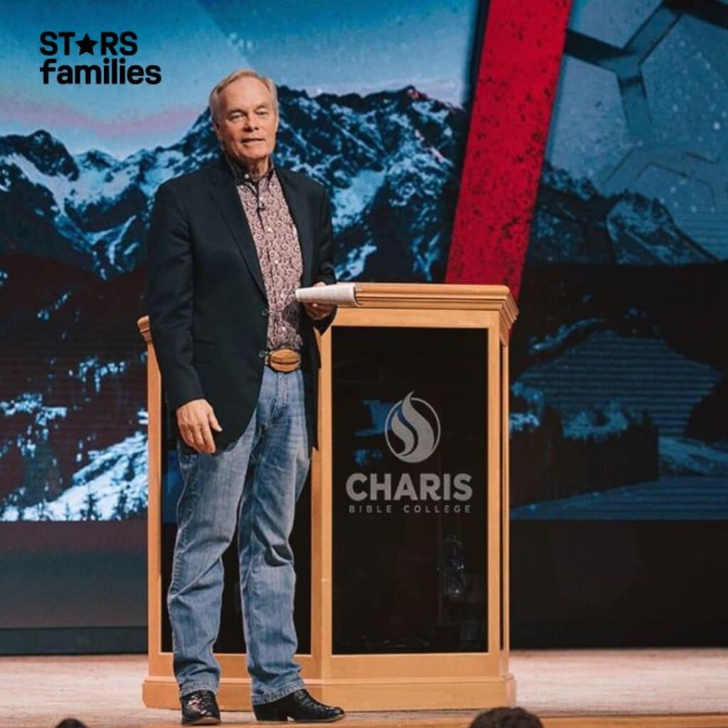 Andrew Wommack, wearing a dark blazer, patterned shirt, jeans, and black shoes, stands behind a podium on a stage. The podium has the logo and name "Charis Bible College" on it. The background features a large screen displaying a mountainous landscape with snow-covered peaks and a red diagonal stripe.