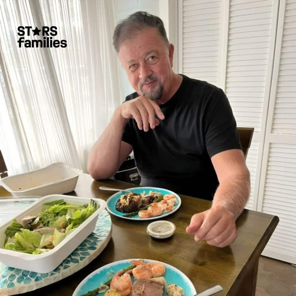 Robert Lichfield, wearing a black shirt, sits at a dining table with two plates of food in front of him. The plate closest to the camera contains shrimp, a piece of meat, mashed potatoes, and some vegetables. The other plate also contains shrimp, a piece of meat, and some vegetables. There is a large bowl of salad on the table, along with small containers of sauce or dressing near each plate.