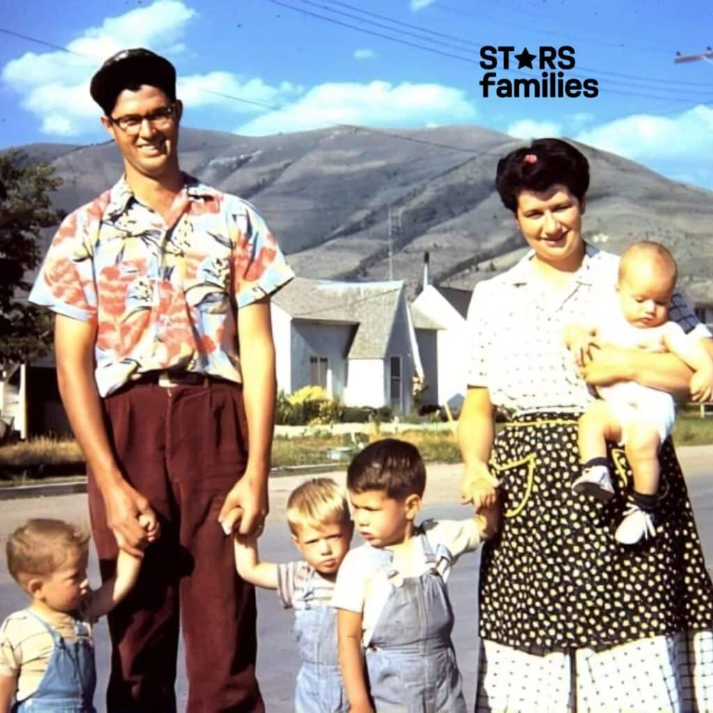 In an old family photograph, Robert Lichfield and his family stand on a street with mountains and houses in the background. Robert Lichfield is wearing a short-sleeved, patterned shirt and dark pants. His wife, in a polka-dotted skirt and a light-colored blouse, holds a baby. Their children, dressed casually, stand with them, with two children holding hands with the parents.