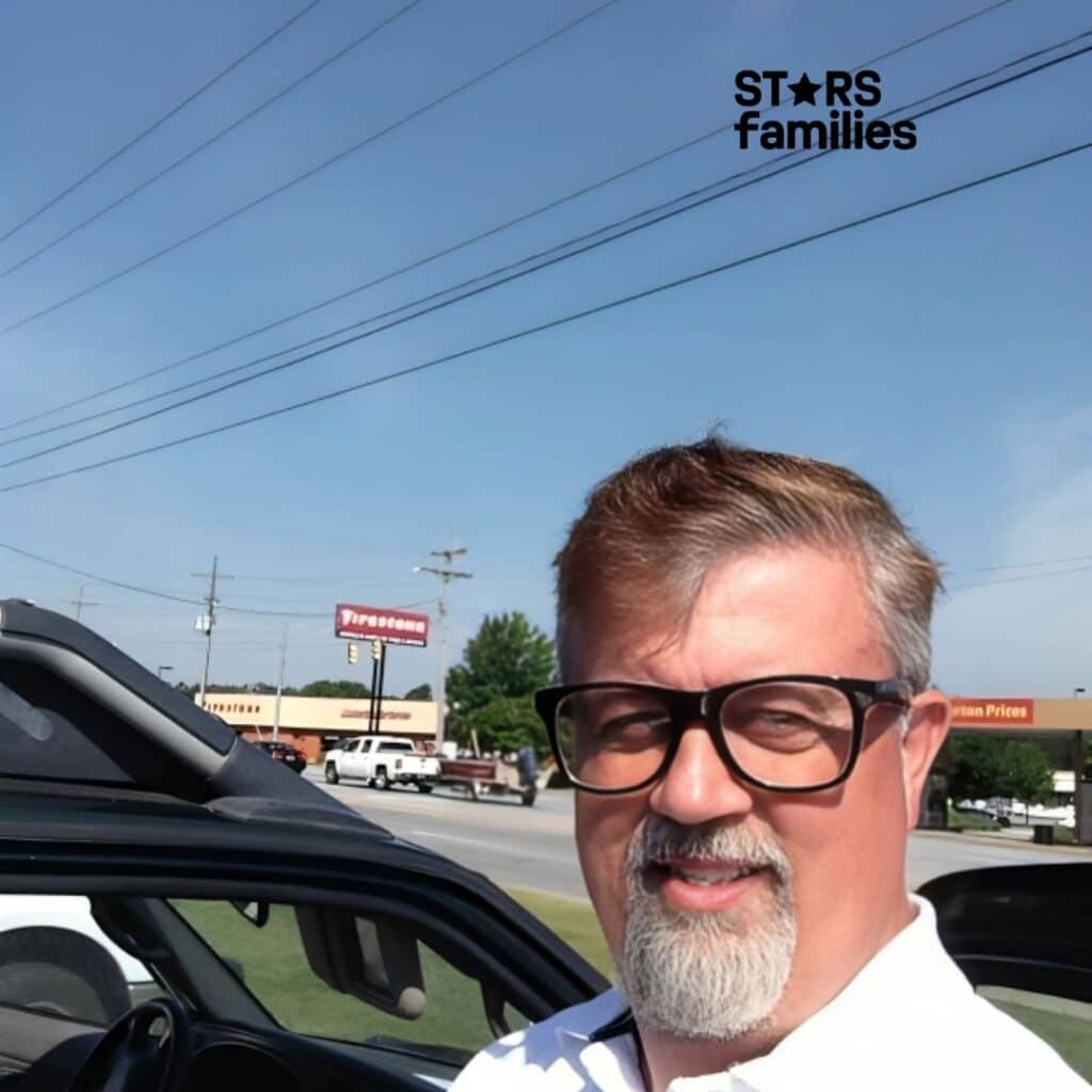 In an outdoor setting, Robert Lichfield is dressed in a white shirt and stands next to an open car door. The background features power lines, a clear blue sky, and several buildings, such as a Firestone store and another building with a sign that says "Urban Prices." There are also trees and a few vehicles on the road.