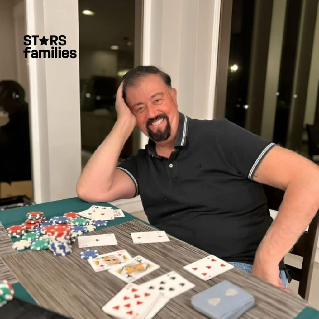 In an indoor setting with large windows showing a night view, Robert Lichfield, dressed in a black polo shirt with white trim on the sleeves and collar, sits at a table covered with poker chips and playing cards. The table features a green tablecloth and a gray placemat, with a deck of cards to the right and a black card shuffler or holder on the left.