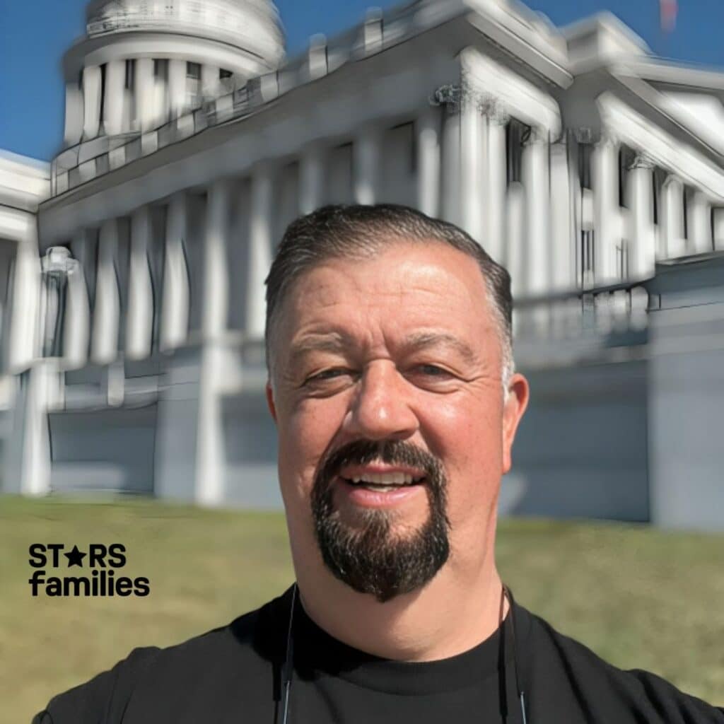 In front of a classical-style building with columns and a dome, Robert Lichfield is dressed in a black shirt with a yellow circular design and has sunglasses hanging around his neck. The building looks like a significant public or government building, such as a capitol building.