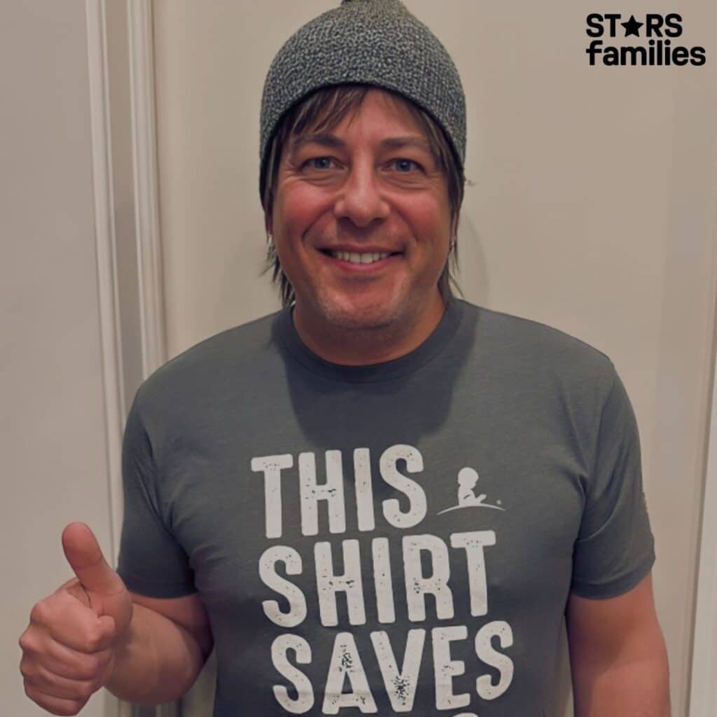 Paul Sidoti, wearing a gray T-shirt with the text "THIS SHIRT SAVES" and a small St. Jude Children's Research Hospital logo, gives a thumbs-up gesture with his left hand. He also wears a knitted beanie. The image promotes a charitable cause supporting St. Jude Children's Research Hospital.