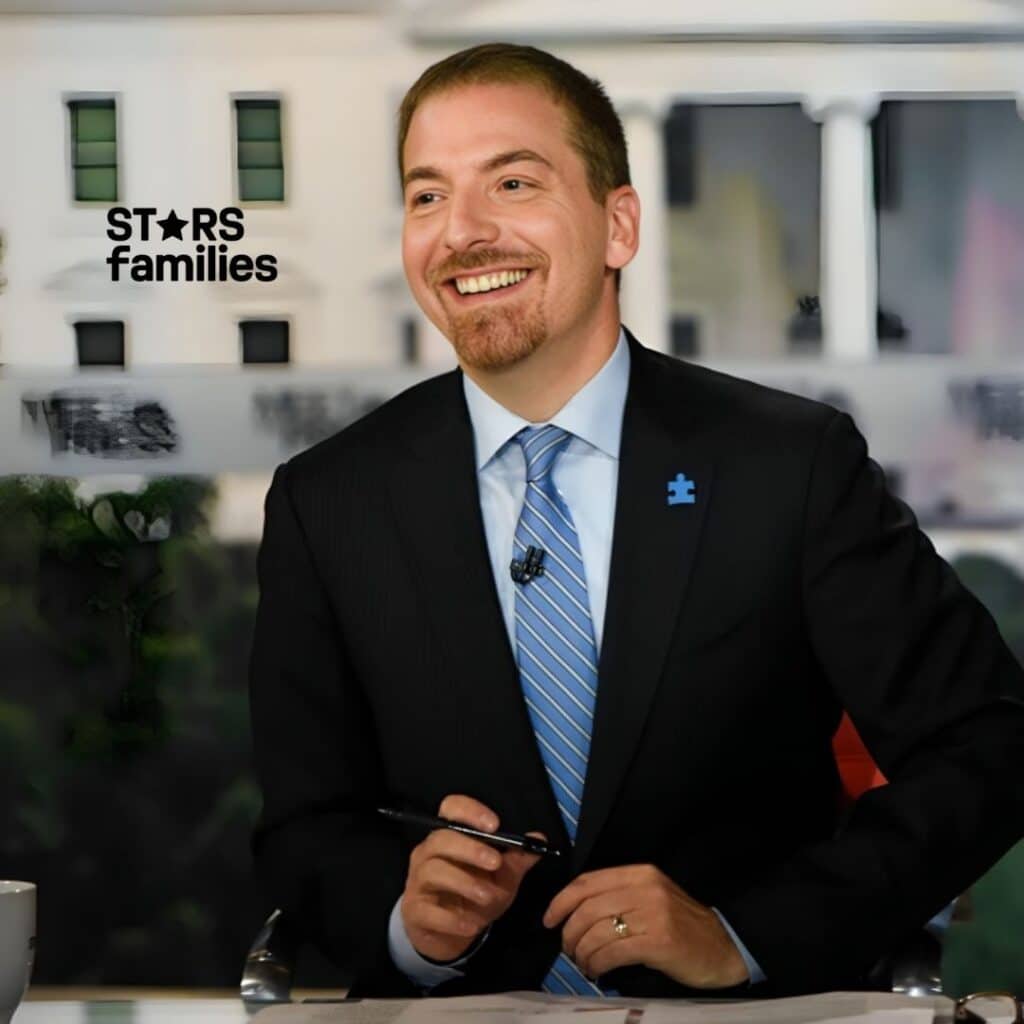 Chuck Todd, wearing a dark suit with a light blue dress shirt and a blue striped tie, is seated at a table holding a pen in his right hand. He has a microphone clipped to his tie and a blue puzzle piece pin on the left lapel of his suit jacket. The background appears to be an out-of-focus image of a building with white walls and green windows, possibly the White House.