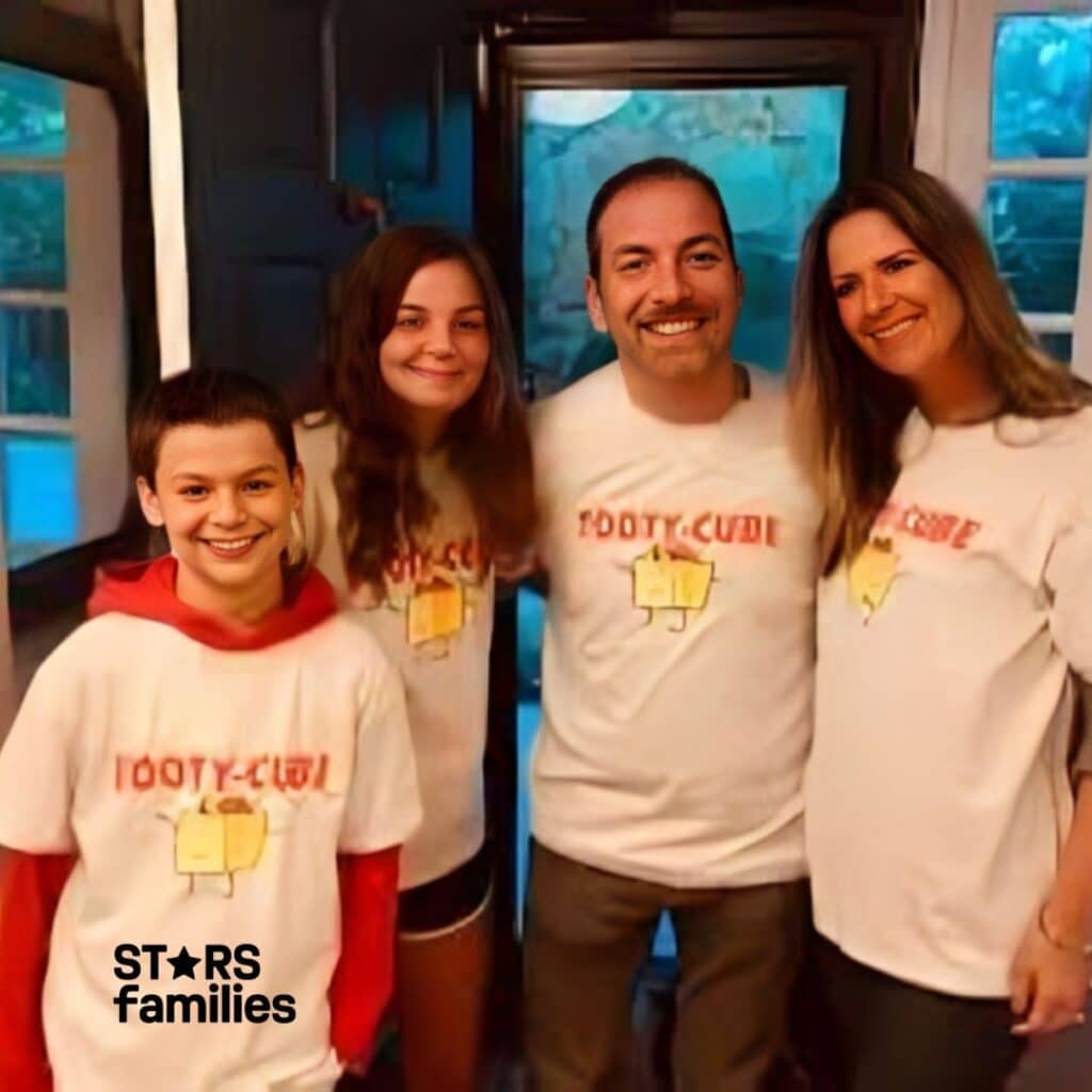 In a home setting, Chuck Todd is dressed in a white T-shirt with a cartoon yellow cube with legs and the text "TOOTY-CUBE" on it. He stands with his wife and two children, who are also wearing matching T-shirts with the same cartoon image and text. The background features a doorway and windows, suggesting they are in a house.