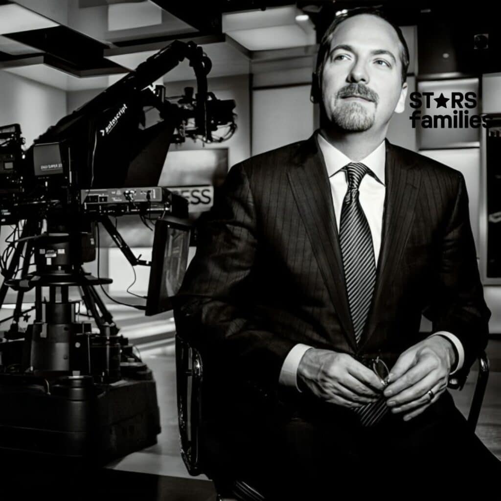 Chuck Todd, wearing a dark pinstripe suit, a white shirt, and a striped tie, is seated in a television studio. The background includes a large camera on a tripod, lighting equipment, and a backdrop with the word "PRESS" partially visible.