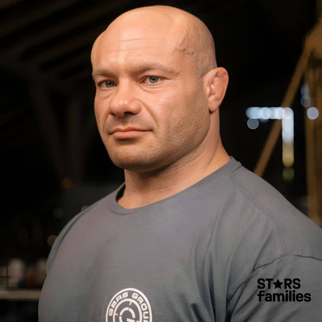 : Mike Israetel, wearing a gray t-shirt with a logo on the left side of the chest. The logo consists of a circular design with the text "GBAS GROUP EST. 2019" surrounding a stylized "G" in the center.