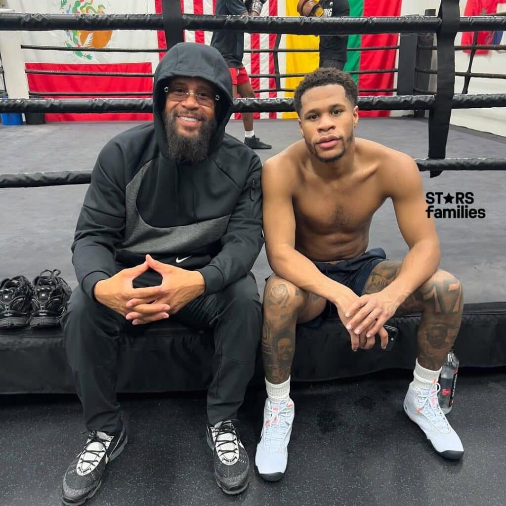 Bill Haney and his son (Devin Haney) sit on the edge of a boxing ring. Bill, wearing a black and gray Nike tracksuit with the hood up, sits on the left. His son, shirtless and wearing black shorts, white socks, and white sneakers, sits on the right.