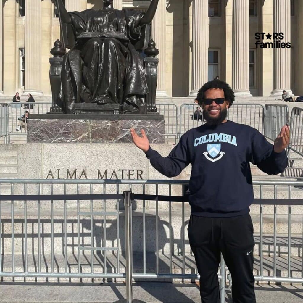 Bill Haney, wearing a navy blue sweatshirt with the word "COLUMBIA" and the university's emblem on it, stands in front of the Alma Mater statue at Columbia University. The statue is a bronze sculpture of a seated female figure, symbolizing wisdom and learning.