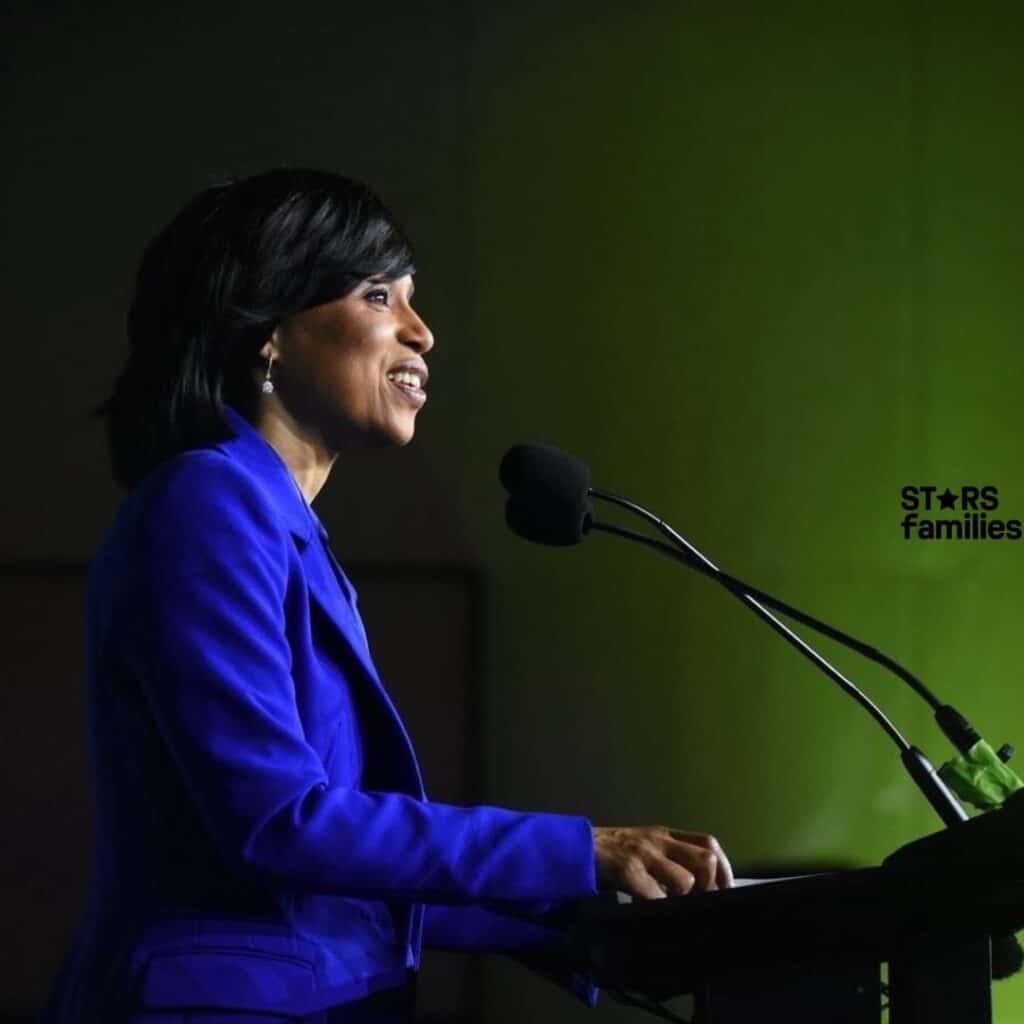 Bill Haney, dressed in a black and gray Nike tracksuit with the hood up, stands on the left, holding a microphone in his left hand. Angela Alsobrooks, wearing a bright blue jacket, stands at a podium speaking into a microphone.