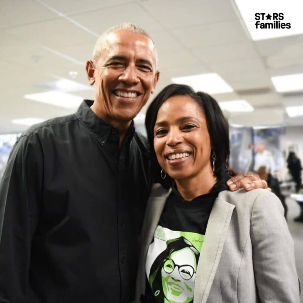 In an indoor setting that appears to be an office or conference room, Barack Obama, dressed in a black shirt, stands beside Angela Alsobrooks, who is wearing a light-colored blazer over a black shirt with a green and white graphic design.