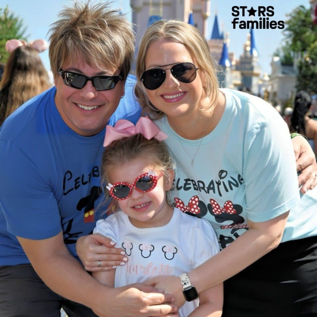 Paul Sidoti, wearing a blue shirt with the text "Celebrating" and a Mickey Mouse design, stands with his wife Ashley and daughter Scarlett in front of a castle at a theme park. Ashley is wearing a light blue shirt with the same text and design, while Scarlett wears a white shirt with a Minnie Mouse design and a pink bow in her hair.