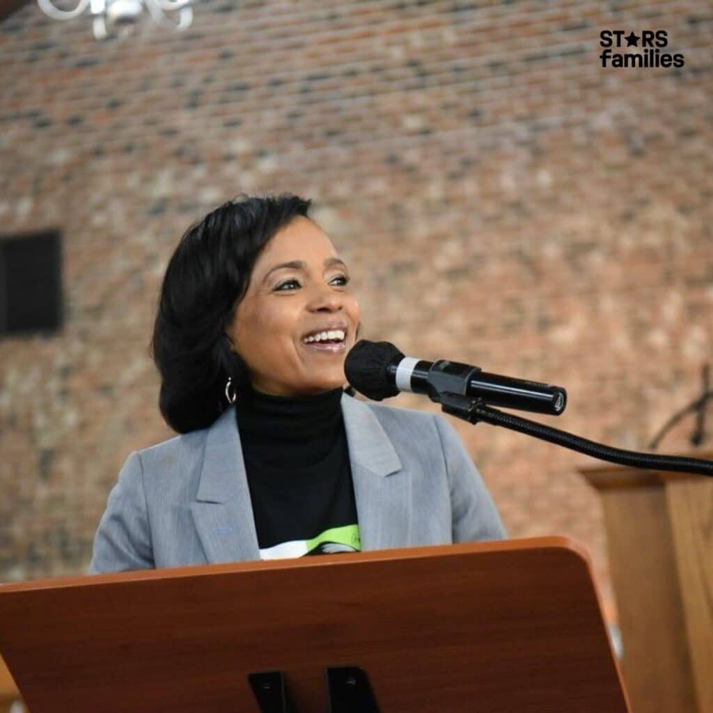 Angela Alsobrooks, wearing a light gray blazer over a black top, stands in front of a brick wall.