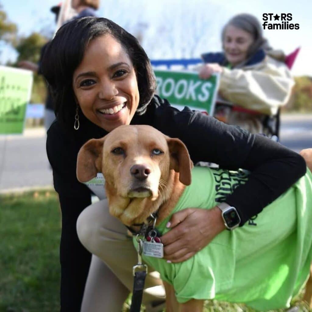 In an outdoor setting, Angela Alsobrooks, dressed in a black long-sleeve shirt and a smartwatch, is crouching down and hugging a dog in a green shirt.