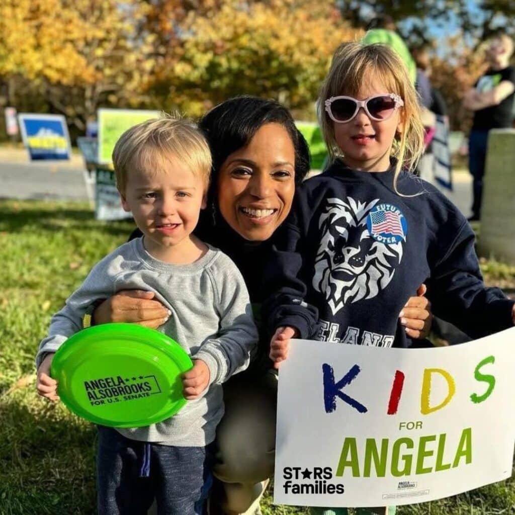 Angela Alsobrooks, wearing a dark outfit, stands outdoors on a grassy area with two children. The child on the left holds a green frisbee with the text "ANGELA ALSOBROOKS FOR U.S. SENATE" printed on it. The child on the right holds a sign that reads "KIDS FOR ANGELA" and wears a sweatshirt with a lion graphic and a "FUTURE VOTER" badge. The background includes trees and gives a sense of a campaign event or rally.