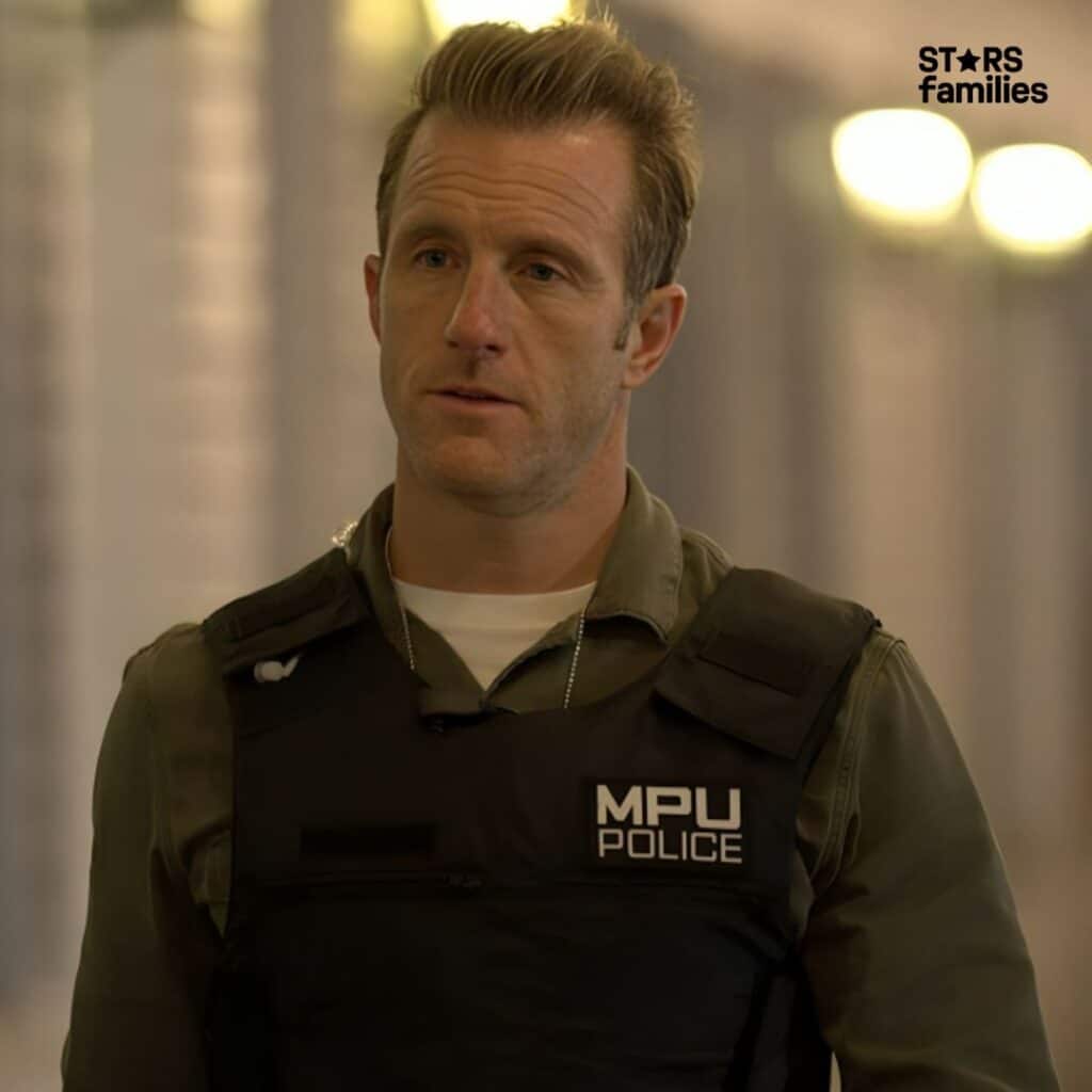 Scott Caan, wearing a police uniform with a vest that has "MPU POLICE" written on it, stands in a corridor or hallway with blurred lights in the background.