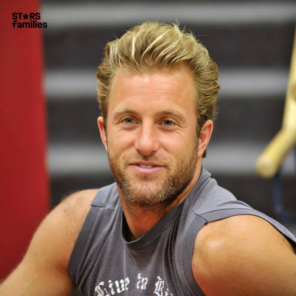 Scott Caan, with blonde hair, is wearing a sleeveless gray shirt with some partially obscured text on it. He is sitting in front of a set of stairs with a red wall on the left side.