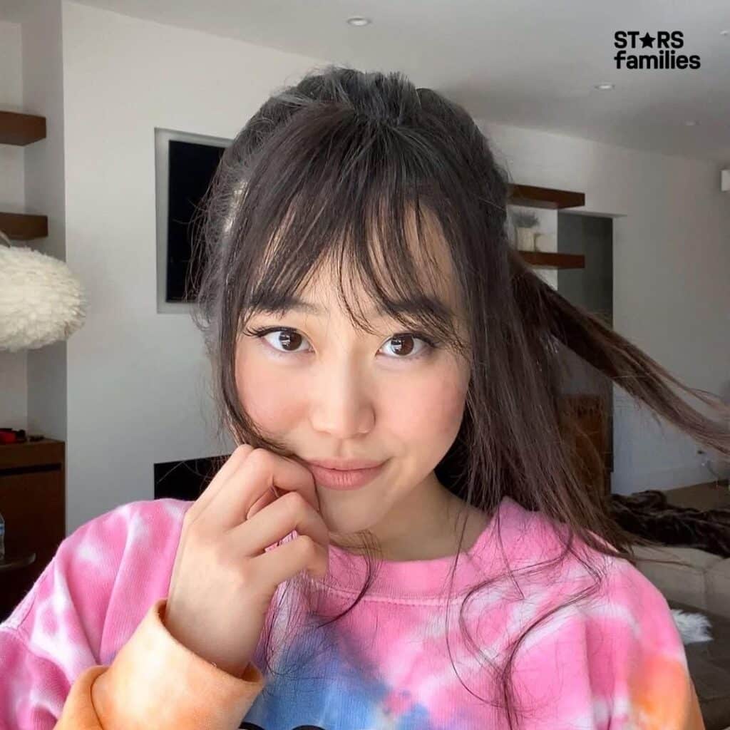 Stephanie Soo, wearing a tie-dye sweatshirt with shades of pink, orange, and blue, holds a hand near her mouth. The background features a modern living room with a white wall, a television mounted on the wall, and some shelves.