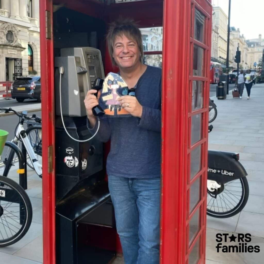 Paul Sidoti stands inside a traditional red British telephone booth, holding a telephone receiver in one hand and a colorful paper cutout in the other hand. The telephone booth contains a payphone. Outside the booth, there are bicycles parked, and a street scene with buildings and pedestrians is visible.