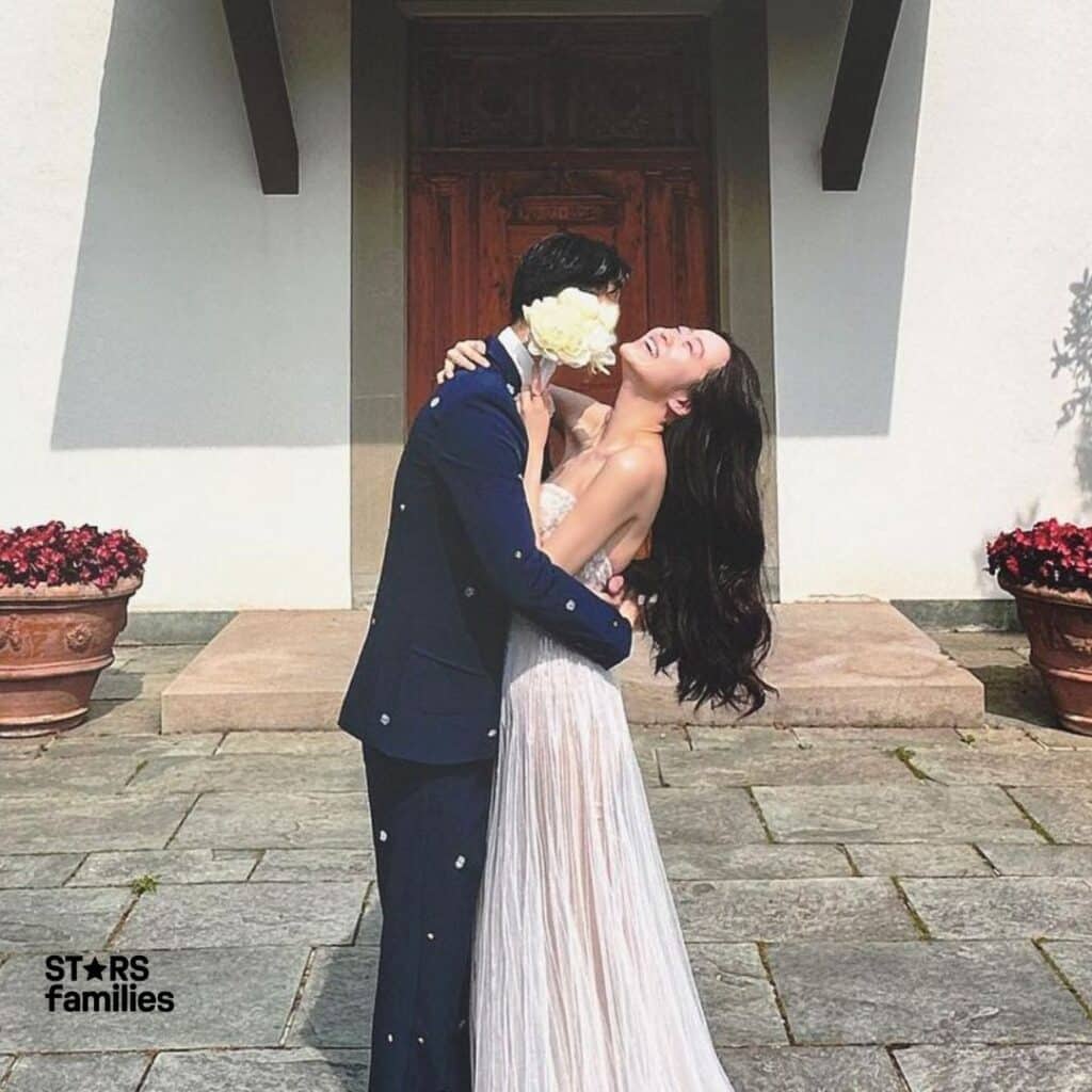 Stephanie Soo, wearing a flowing strapless white gown, stands next to her husband, who is dressed in a dark blue suit adorned with small decorative elements. They are standing in front of a large wooden door with potted plants on either side, suggesting an outdoor setting. The image captures a significant and intimate moment between the couple, possibly from their wedding or a similar special occasion.