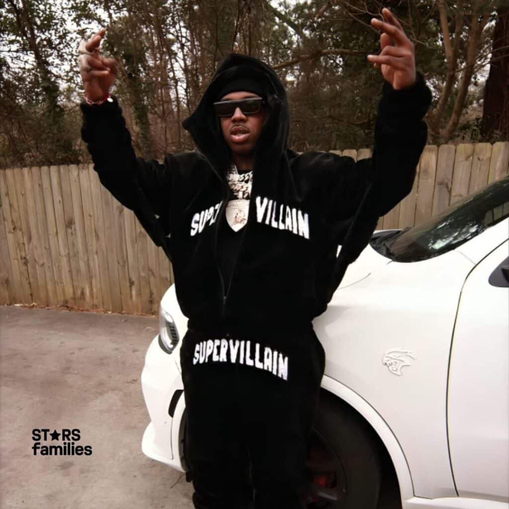 EST Gee, wearing a black hoodie and matching pants with the word "SUPERVILLAIN" written in white letters, stands in front of a white car with a "Hellcat" logo on the fender. He is wearing white sneakers and has a chain around his neck. His face is blurred for privacy. The background includes a wooden fence and trees.