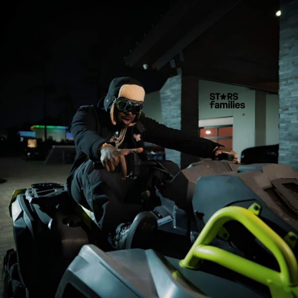 In a nighttime setting, EST Gee, dressed in a black hoodie with multiple chains around the neck, poses on an ATV.