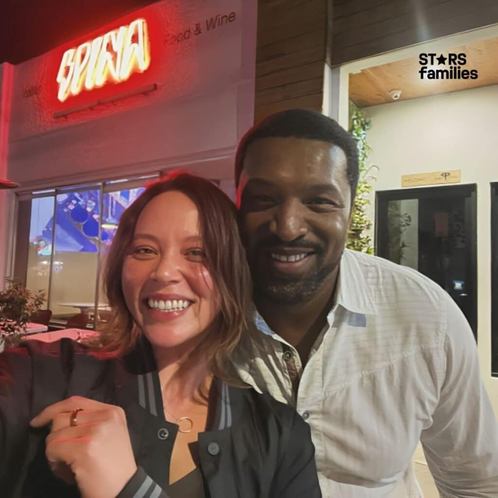 In front of a restaurant with a neon sign reading "SPIGA" and "Food & Wine," Melissa O'Neil, dressed in a black dress, stands beside another person.