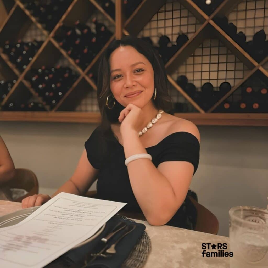 In a restaurant or wine bar setting, Melissa O'Neil, dressed in a black off-shoulder top, a pearl necklace, a white bracelet, and gold hoop earrings, sits at a table holding a menu with one hand while resting her chin on the other hand. The table has a glass of water and a set of cutlery on a napkin. The background includes a wine rack filled with wine bottles.