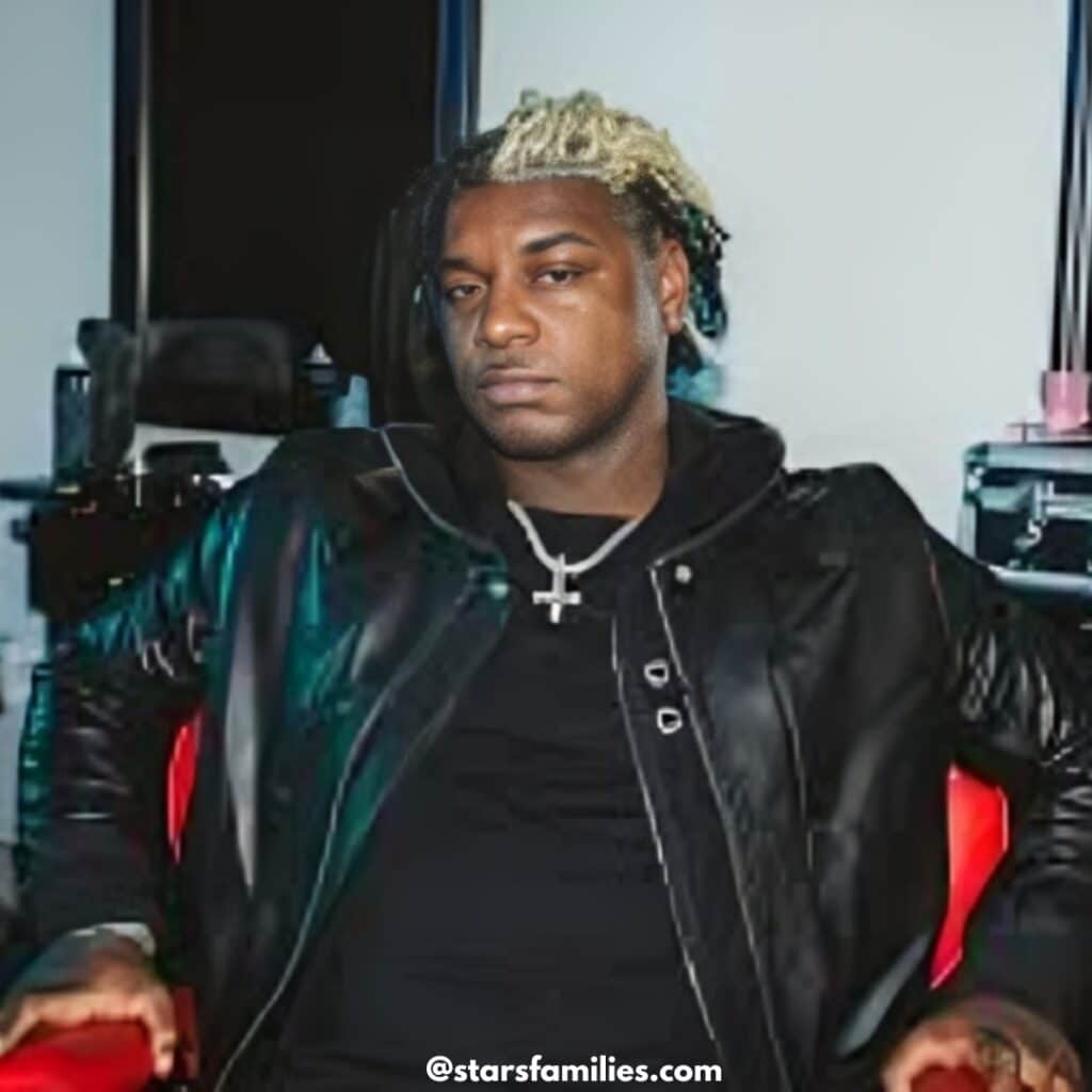 Ken Carson, dressed in a black leather jacket over a black shirt, sits on a red chair. He is wearing a necklace with a cross pendant.