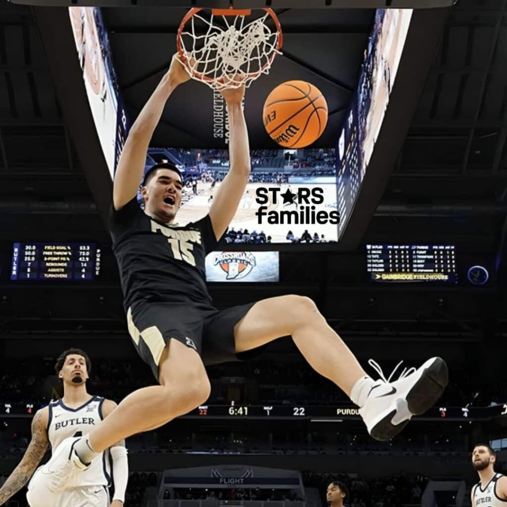 Zach Edey, wearing a black Purdue jersey with the number 15, performs a powerful slam dunk, hanging onto the rim with both hands. In the background, a player from Butler in a white jersey watches the play.