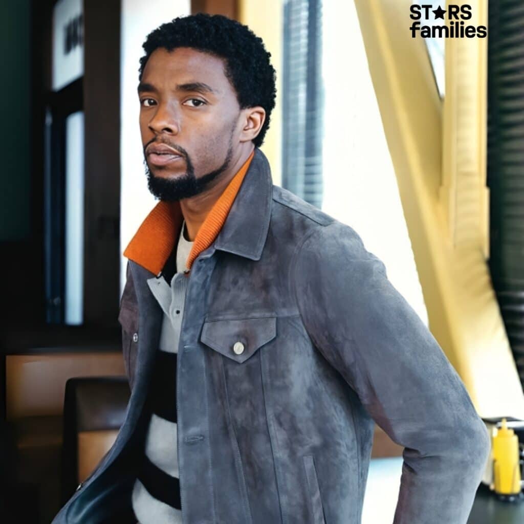 In a casual indoor setting, Chadwick Boseman sports a gray suede jacket over a striped sweater with black, white, and gray horizontal stripes. He is standing next to a window with blinds and a yellow structural element.