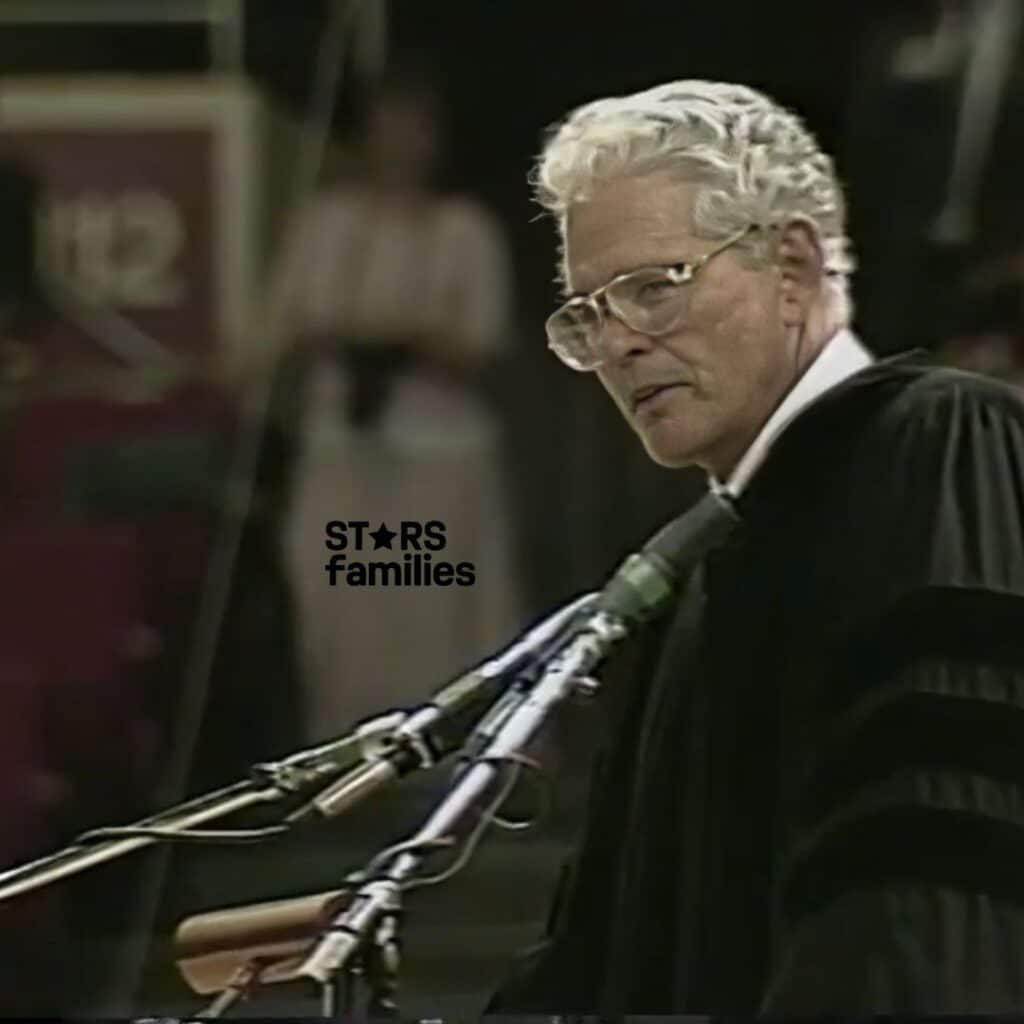 In an auditorium setting, John Teets, dressed in a black academic gown with velvet stripes, speaks at a podium with multiple microphones.