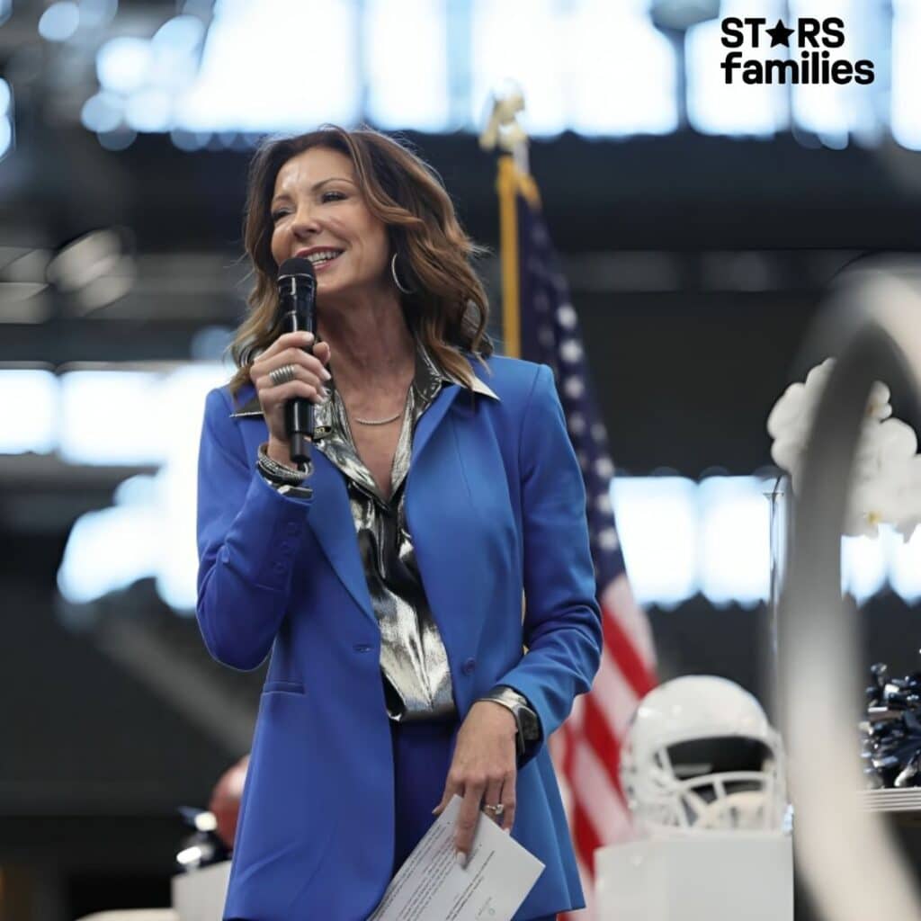 Charlotte Jones, wearing a blue blazer over a metallic silver shirt, speaks into a microphone at an event. The background includes an American flag and a football helmet. She is holding a piece of paper with text on it.