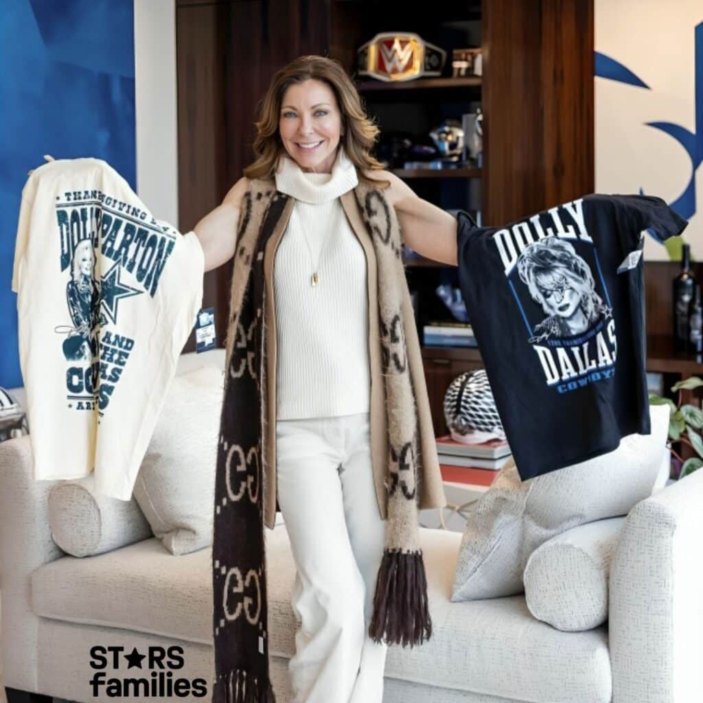 Charlotte Jones stands in a room, holding two t-shirts. The t-shirt on the left is white with text that reads "THANKSGIVING DOLLY PARTON AND THE COWBOYS." The t-shirt on the right is black with text that reads "DOLLY DALLAS COWBOYS." She is wearing a white outfit with a long scarf.