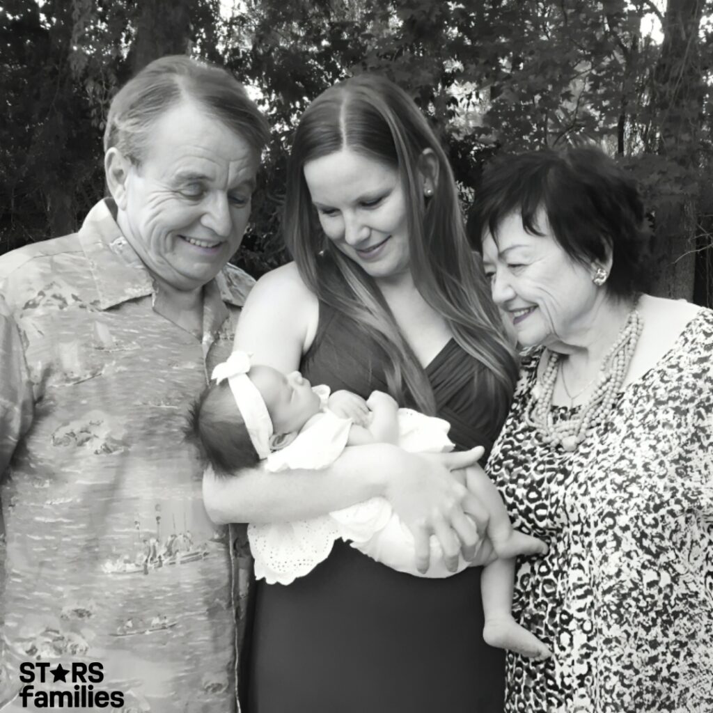 Four generations of a family pose together outdoors. in the picture his mother and daughter holding a baby dressed in a light-colored outfit with a headband.