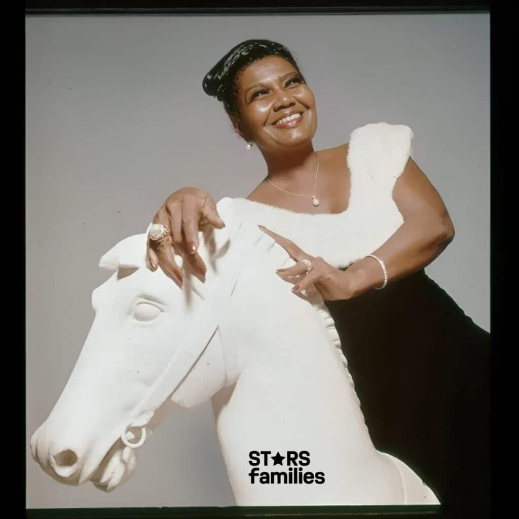 Pearl Bailey, dressed in an elegant outfit with intricate jewelry, poses with her husband beside a white sculpture of a horse's head.