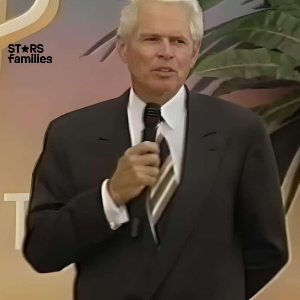 John Teets, wearing a suit and holding a microphone, stands in front of a backdrop with the word "CERT" visible as part of a larger word or phrase. A large plant is also in the background.